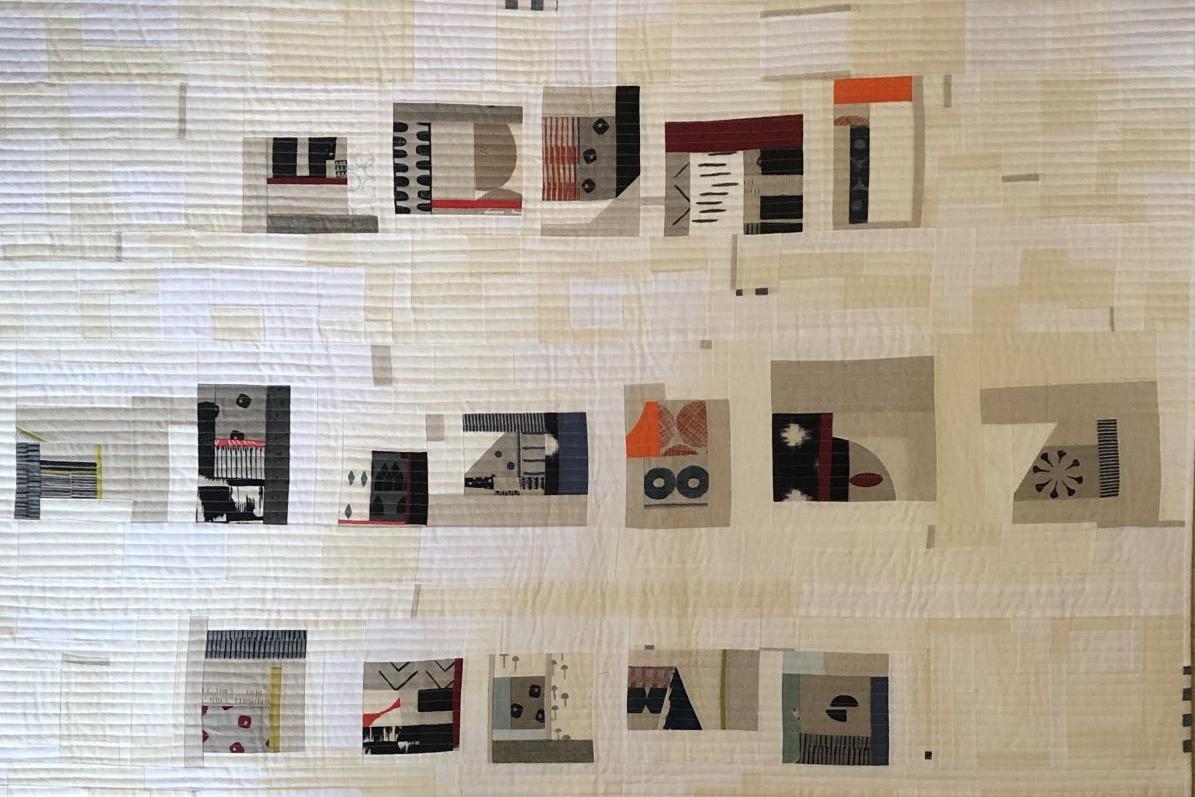 A large, abstract quilt with a predominantly white background. The quilt is composed of various shapes and patterns in black, gray, and pops of color like red and orange. There are geometric shapes, organic forms, and textural elements, creating a complex