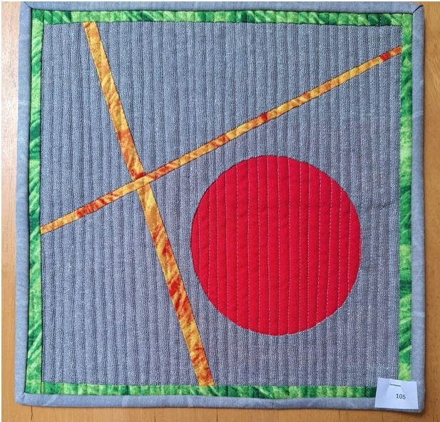 A square quilt with a gray background and a green border. The quilt features a large, red circle and two yellow lines intersecting it. The background is quilted with a linear pattern.