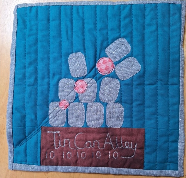 A square quilt with a blue background and a gray border. The quilt features a stack of gray cans with red balls on top, resembling a game of Tin Can Alley. The phrase "Tin Can Alley" is embroidered at the bottom. The background is quilted with a linear pattern.