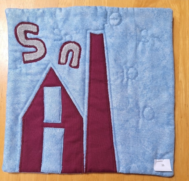 A square quilt with a blue background and a red border. The quilt features a red house with a chimney and the letters "Sn" in gray.
