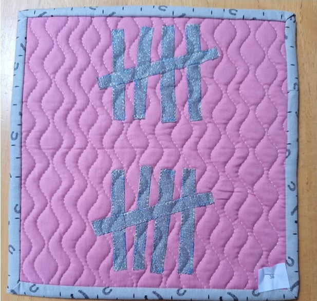 A square quilt with a pink background and a gray border. The quilt features two silver hashtag symbols on the pink background. The background is quilted with a wavy pattern.