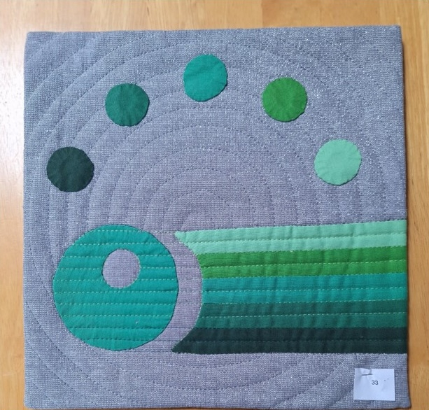 A square quilt with a gray background and a green border. The quilt features a series of green circles arranged in a semi-circle, with a larger circle in the centre. There is also a horizontal strip of green fabric with varying shades of green.