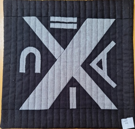 A square quilt with a black background. The quilt features a geometric design with intersecting lines and shapes, including a triangle, a circle, and a rectangle. The shapes are made up of silver fabric and are quilted with a linear pattern.