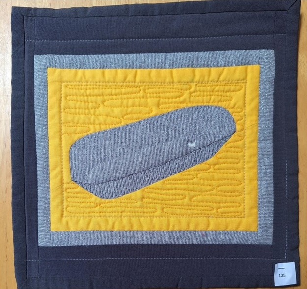 A square quilt with a dark grey background and a yellow centre. The quilt features a grey, rectangular tin bath, placed on a yellow background.