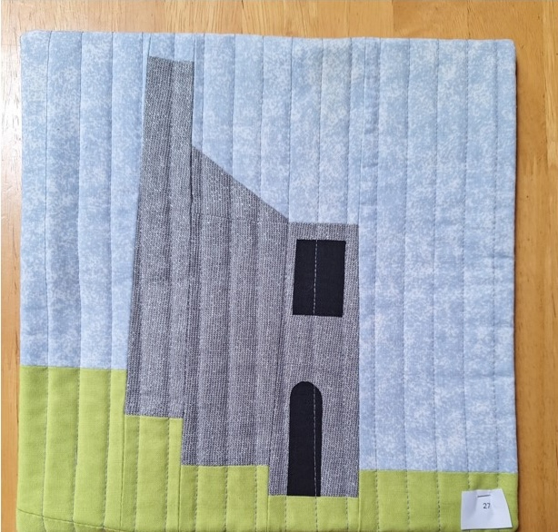 A square quilt with a gray background and a green border. The quilt features a gray building with a window and a door, standing on a green patch of ground. The background is quilted with vertical lines.