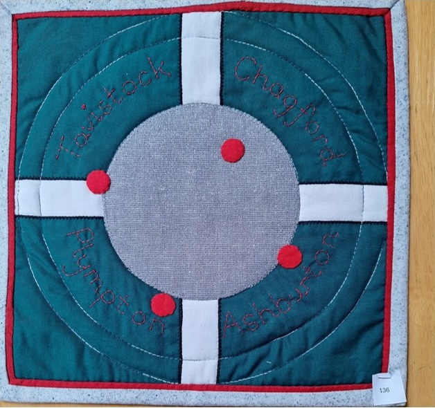 A square quilt with a green background and a red border. The quilt features a gray circle in the centre with the word "Tin" written around the edge. There are four red circles around the gray circle, and the background is quilted with a circular pattern.
