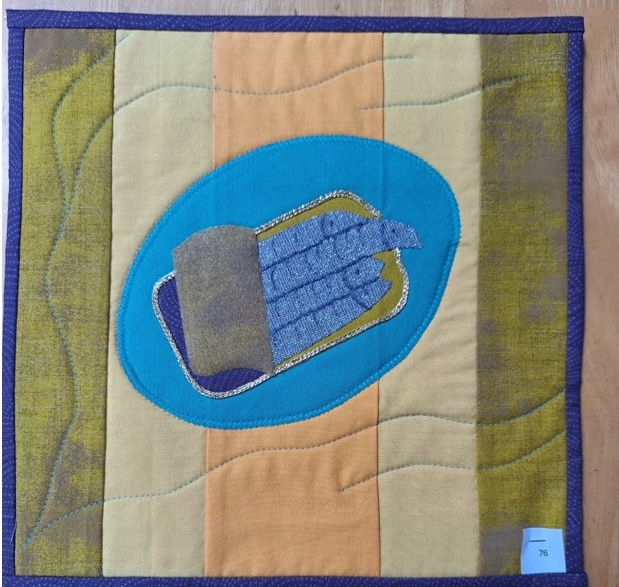 A square quilt with a blue background and a purple border. The quilt features a blue oval shape representing a tin can, with a silver lid and several sardines inside. The background is a patchwork of green and orange fabric with a wavy pattern