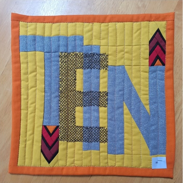 A square quilt with a yellow background and an orange border. The word "TEN" is spelled out in gray letters, with the letter "E" having a textured, patterned design. The background is quilted with vertical lines.