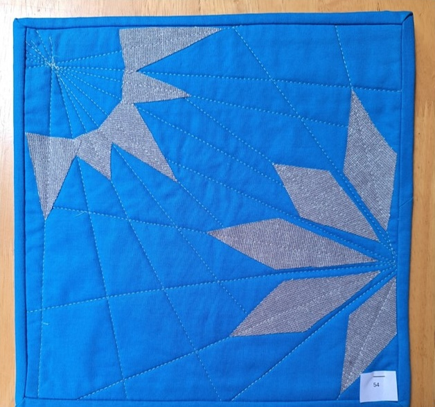 A square quilt with a blue background and a blue border. The quilt features a geometric design with overlapping triangles in gray, creating a star-like shape. The background is quilted with a linear pattern