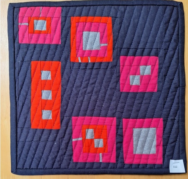 A square quilt with a dark blue background. The quilt features a pattern of pink and red squares, some of which contain smaller squares within them. The background is quilted with a diagonal line pattern.