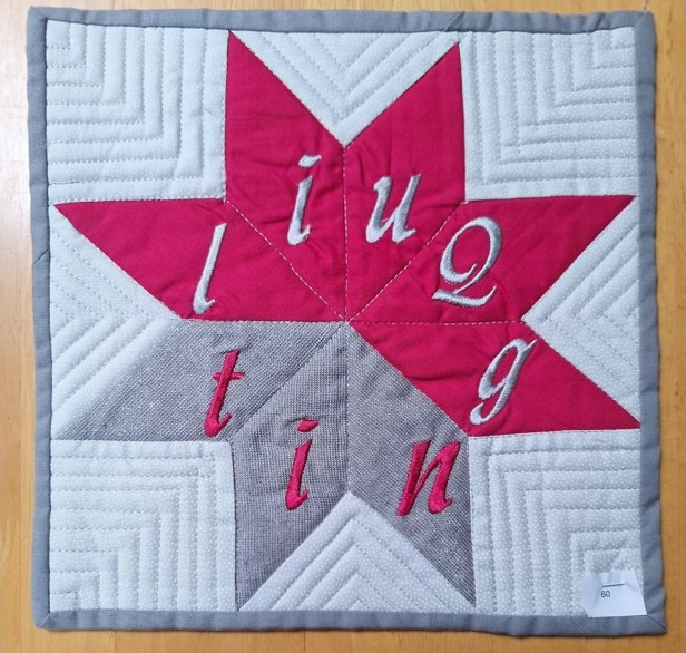 A square quilt with a red and gray star shape in the centre. The letters "i", "u", "q", "g", "t", "i", and "n" are embroidered on the star points. The background is quilted with a linear pattern.