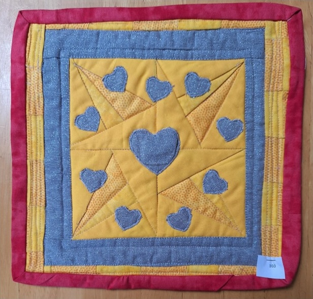A square quilt with a yellow background and a red border. The quilt features a star-shaped design made up of smaller triangles, with grey heart shapes placed in the centre of each triangle.