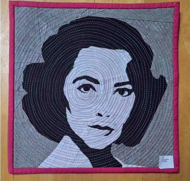 Square quilt with a monochrome appliqué of a woman as the focal point. Circular quilting radiates out from her eye in bright colours. Hot pink binding finishes the quilt edges.