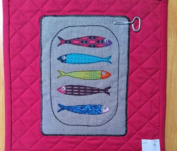 A square quilt with a red background. The quilt features a tin of sardines with five colourful sardines inside. A small key is visible on the top of the tin. The background is quilted with a hatched pattern.