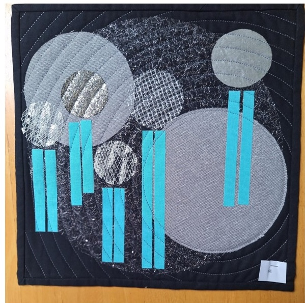 A square quilt with a black background and a black border. The quilt features overlapping circles in gray, with some having textured surfaces. Blue vertical lines intersect the circles. The background is quilted with a circular pattern.