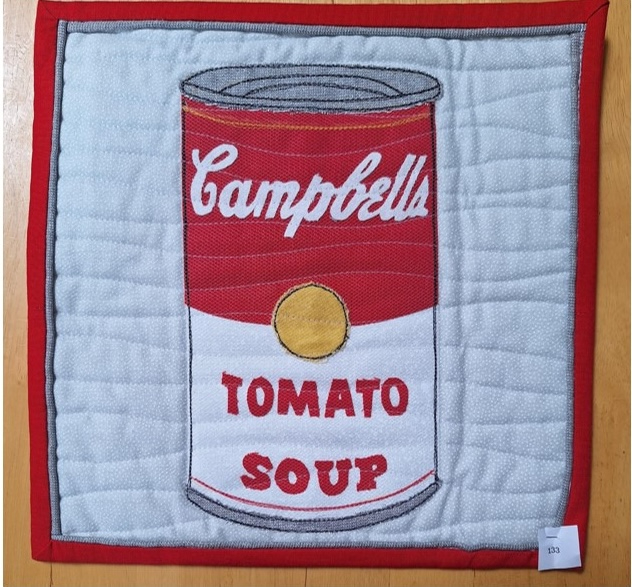 A square quilt with a red border. The quilt features a Campbell's Tomato Soup can, depicted with a red background and white lettering. The background is quilted with a wavy line pattern.