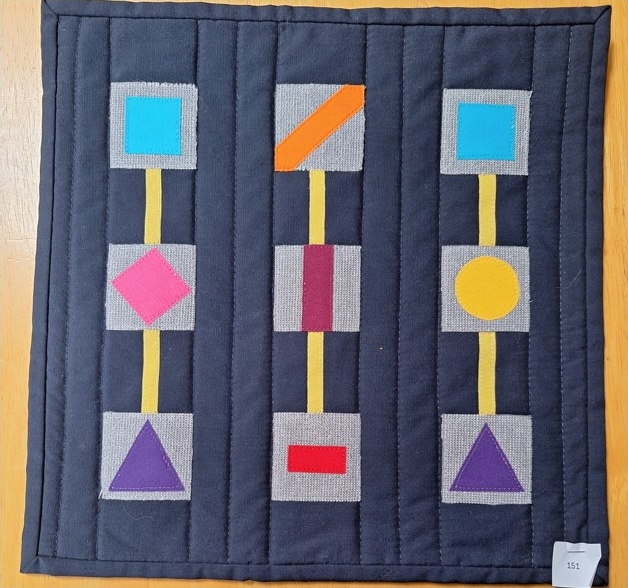 A square quilt with a black background. The quilt features three vertical columns of colourful shapes, including squares, rectangles, triangles, circles, and diamonds. The shapes are connected by yellow lines. The background is quilted with a linear pattern.