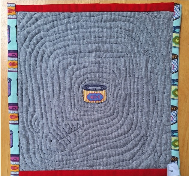 A square quilt with a gray background and a red and blue border. The quilt features a small, colourful can in the centre, surrounded by a swirling, quilted pattern. The background is quilted with concentric circles which hides a fish skeleton.