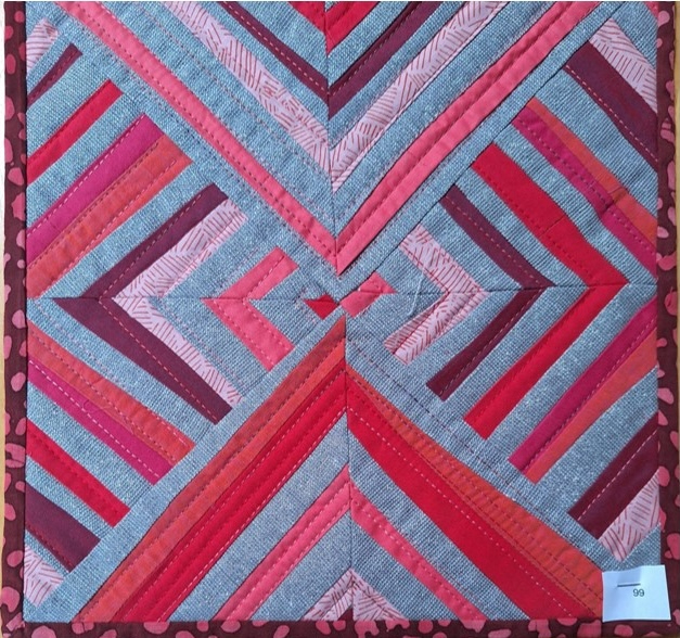 A square quilt with a gray background and a pink border. The quilt features a geometric design with intersecting lines and shapes in various shades of pink and red.