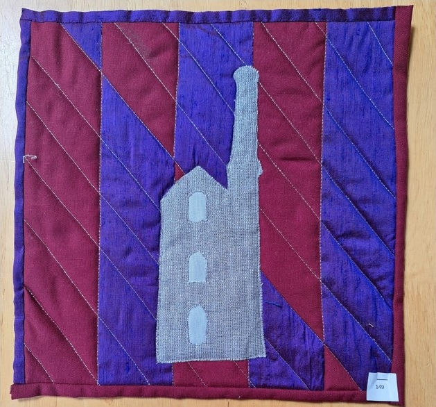 A square quilt with a red and purple background. The quilt features a gray silhouette of a tin mine with a tall chimney. The background is quilted with a diagonal line pattern.