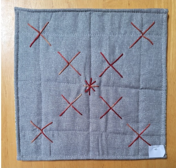 A square quilt with a gray background and a red border. The quilt features a pattern of red "X" shapes, with a smaller "X" in the centre.