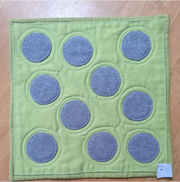 A square quilt with a green background and a green border. The quilt features nine gray circles arranged in a grid-like pattern.