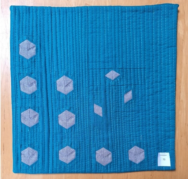 A square quilt with a blue background. The quilt features multiple gray, hexagonal shapes of varying sizes, arranged in a staggered pattern. The background is quilted with a linear pattern.