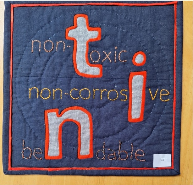 A square quilt with a dark blue background and a red border. The quilt features the words "non-toxic" and "non-corrosive" in large, white, embroidered letters. The words "be" and "able" are embroidered in smaller, white letters below.