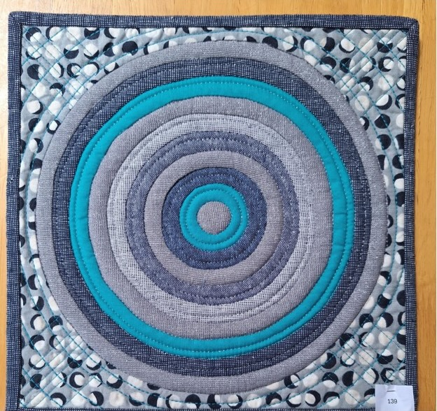 A square quilt with a gray background and a black border. The quilt features a series of concentric circles in shades of gray and turquoise. The background is quilted with radiating circles