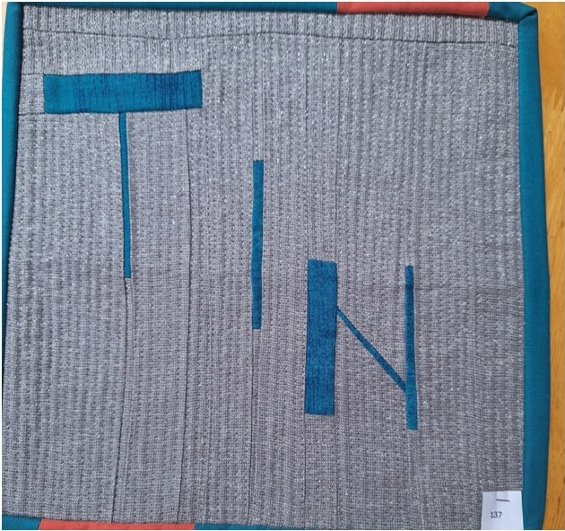 A square quilt with a gray background and a red border. The word "TIN" is spelled out in blue letters, with each letter made up of vertical and horizontal lines. The background is quilted with a linear pattern.