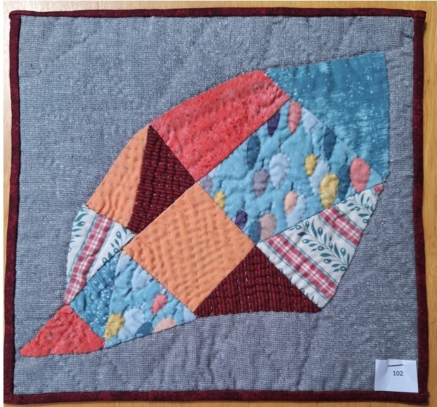 A square quilt with a gray background and a red border. The quilt features a large, abstract shape resembling a leaf or petal, made up of various colourful fabric pieces in shades of red, orange, blue, and green.
