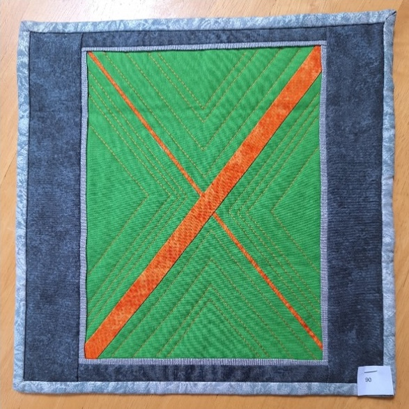 A square quilt with a green background and a gray border. The quilt features two intersecting orange lines forming an "X" shape. The background is quilted with a chevron pattern.