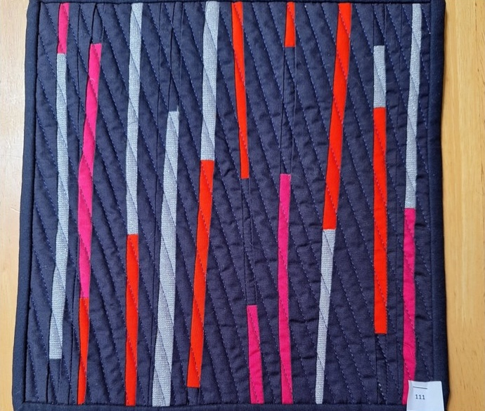 A square quilt with a dark blue background. The quilt features vertical strips of red, pink, and grey fabric, arranged in a staggered pattern. The background is quilted with a diagonal line pattern.