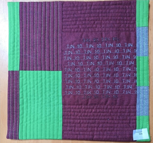 A square quilt with a purple background and a green border. The word "TIN" is repeated multiple times in gray letters, arranged in a grid-like pattern. The background is quilted with vertical lines.