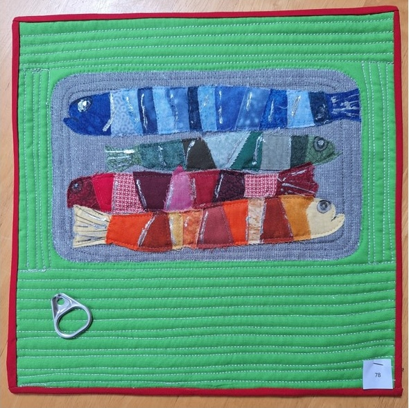 A square quilt with a green background and a red border. The quilt features three colourful fish arranged in a row, with a tin can ring at the bottom. The fish are made of various fabrics with different patterns and textures. The background is quilted with vertical lines.