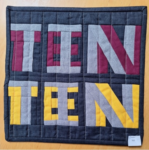 A square quilt with a black background and a black border. The word "TEN" is spelled out twice in large, colourful letters. The first "TEN" is made up of red, gray, and yellow fabric, while the second "TEN" is made up of gray and yellow fabric. The background is quilted with a linear pattern.
