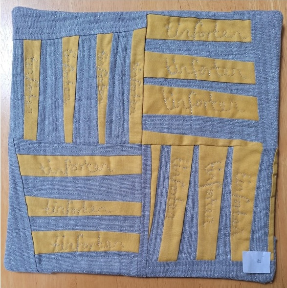 A square quilt with a gray background and a gray border. The quilt features a pattern of yellow rectangles with the word "Winter" embroidered on them. The rectangles are arranged in a grid-like pattern on the gray background. The background is quilted with a linear pattern.