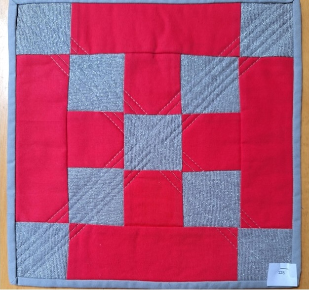 A square quilt with a red background and a gray border. The quilt features a pattern of alternating red and gray squares, with each square having a cross-shaped design stitched onto it. The background is quilted with a linear pattern.