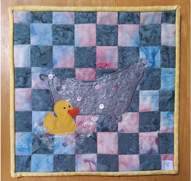 A square quilt with a checkered background in shades of blue and pink. A yellow rubber duck is placed in a bathtub, which is made of a textured, grey fabric. The background is quilted with a linear pattern. The quilt has a yellow border.