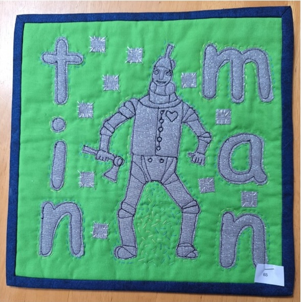 A square quilt with a green background and a blue border. The quilt features the Tin Man from The Wizard of Oz, standing in the centre. The word "TIN MAN" is spelled out in gray letters around him.
