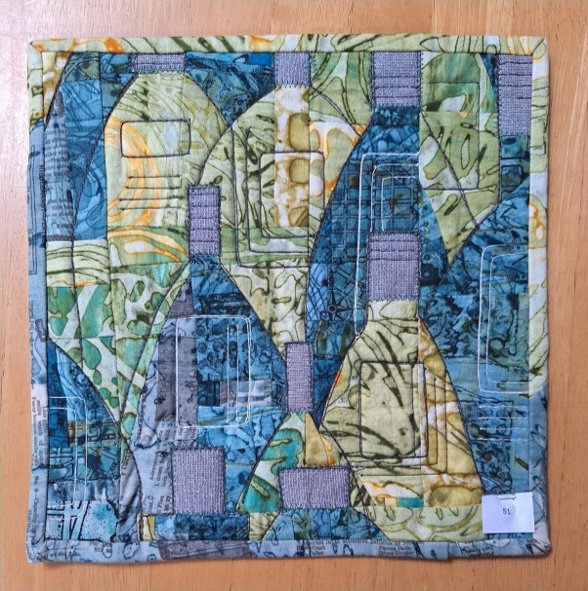A square quilt with a blue background and a gray border. The quilt features a patchwork design with overlapping shapes in blue, green, and gray. The shapes are outlined with white stitching and have various textures and patterns.