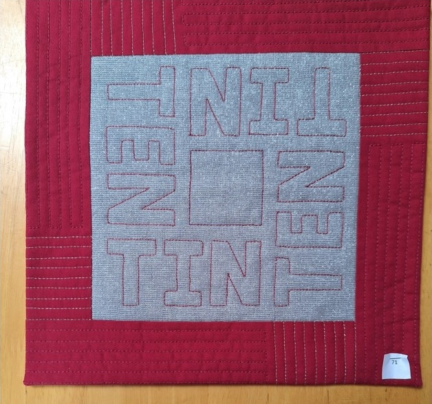 A square quilt with a red background and a red border. The word "TIN" is repeated four times in gray letters, arranged in a square pattern. The background is quilted with a linear pattern.