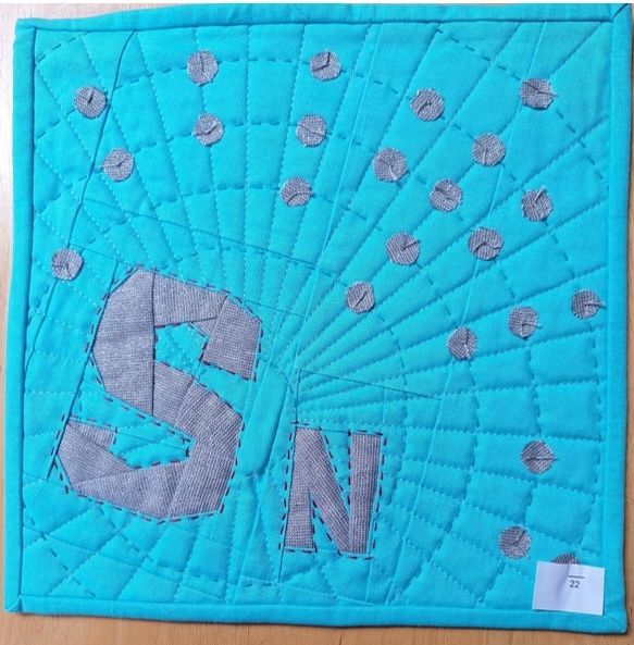 A square quilt with a blue background and a gray border. The quilt features the letter "Sn" in gray, surrounded by a circular pattern of small, gray circles. The background is quilted with a radiating line pattern.