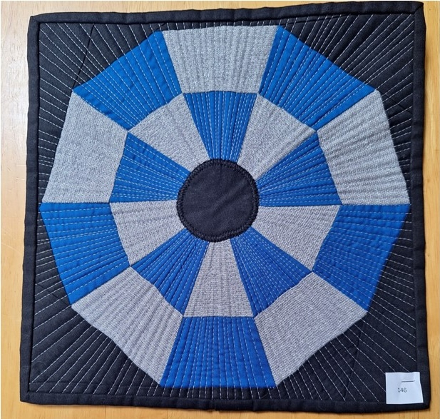A square quilt with a black background and a blue border. The quilt features a circular design with radiating triangles in shades of blue and gray. The centre of the circle is a solid black circle. The background is quilted with straight lines radiating out from the centre.