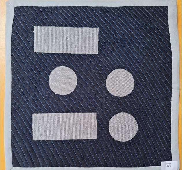 A square quilt with a dark blue background and a gray border. The quilt features a rectangle, two circles, and another rectangle arranged in a vertical pattern. All shapes are gray and the background is quilted with a diagonal line pattern.