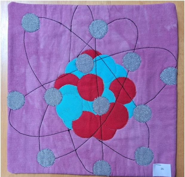 A square quilt with a purple background and a gray border. The quilt features a stylized atom with a blue nucleus and red and gray electrons orbiting around it.