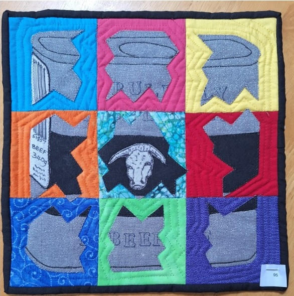 A square quilt with a colourful background and a black border. The quilt features nine squares, each containing a different image related to food and drink, such as a can of soup, a head of cattle, and a bowl of fruit. The background is quilted with a linear echo pattern.