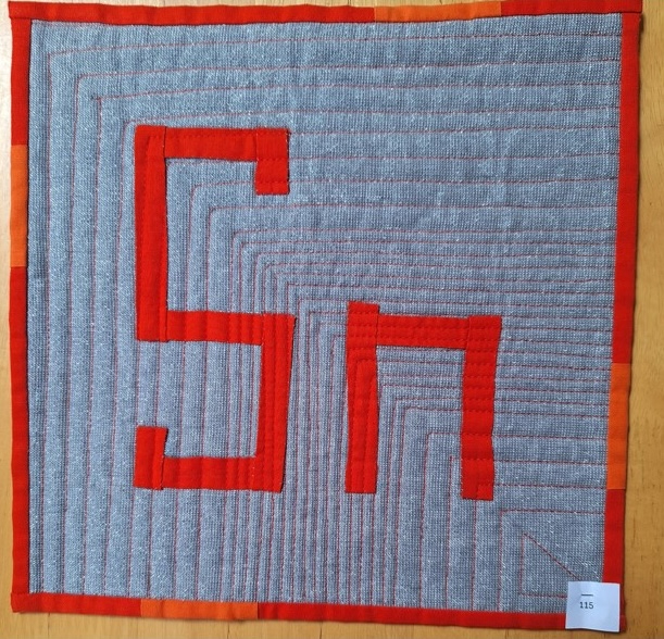 A square quilt with a grey background and an orange border. The quilt features the letters "Sn" made up of red fabric pieces. The background is quilted with a linear pattern.
