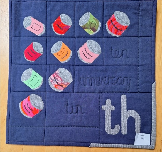 A square quilt with a blue background and a blue border. The quilt features a grid of colourful tin cans, with the words "ten" and "anniversary" and the number "10th" embroidered in the bottom right corner. The background is quilted with a grid-like linear pattern.