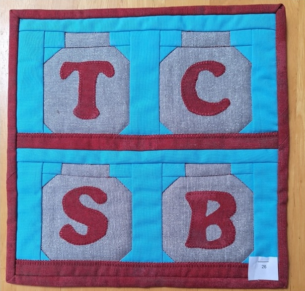 A square quilt with a blue background and a red border. The quilt features four gray squares, each with a letter: "T", "C", "S", and "B" in red.